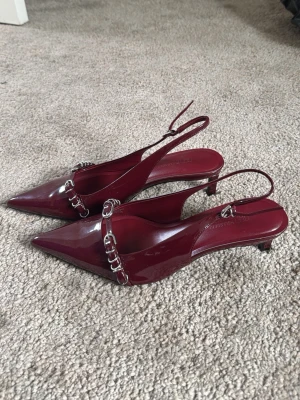 Vinröda lackpumps med kedjedetalj - Säljer ett par snygga vinröda slingback-klackar med spetsig tå och dekorativa spännen framtill. Skorna är köpta från Pull & bear. Endast provade, så de är i mycket fint skick.  * Storlek: 38 * Passar mig som vanligtvis har storlek 38 * Färg: Vinröd/Burgundy * Skick: Endast provade 