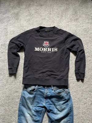Svart sweatshirt från Morris - Stilren svart sweatshirt från Morris med broderad logga och texten 'Morris Since 1968' på bröstet. Tröjan har rund halsringning och ribbade muddar vid ärmslut och nederkant. Bra skick, storlek small.