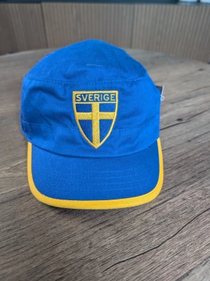 Blå keps med Sverige-emblem - Snygg blå keps med gul kant runt skärmen och broderat Sverige-emblem framtill. Kepsen har klassisk form och ventilationshål på sidorna. Perfekt för att visa ditt stöd för Sverige. Materialet ser ut att vara bomull.
