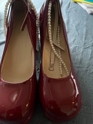 Vinröda lackballerinaskor med pärlband - Säljer ett par vinröda ballerinaskor i glansigt lackmaterial. Skorna har rund tå och dekorativa pärlband samt ett smalt justerbart band över foten. Perfekta för dig som vill ha en söt och unik detalj till din outfit.