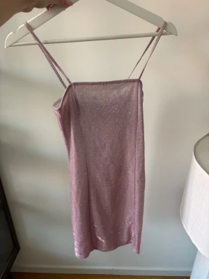 Glittrig rosa kort klänning - Säljer en glittrig rosa kort klänning med smala axelband och rak urringning. Klänningen har en enkel, figurnära passform. Oanvänd med prislapp. 