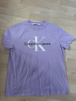 Ljuslila Calvin Klein t-shirt - Säljer en ljuslila t-shirt från Calvin Klein Jeans med stor vit CK-logga och svart text på bröstet. Klassisk rund hals och korta ärmar. Perfekt för dig som gillar stilrena och trendiga plagg.