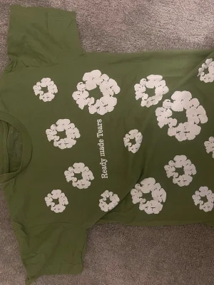 Denim tears tshirt  - Snygg grön denim tears t-shirt med vita blommor tryckta över hela framsidan och texten 'Ready made Tears' i mitten. T-shirten har klassisk passform och rund hals. Perfekt för dig som vill sticka ut med ett coolt och lekfullt tryck.