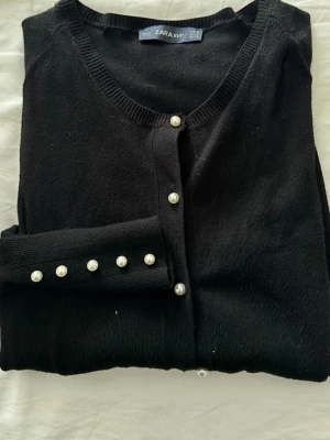 Svart kofta med pärlknappar från Zara - Stilren svart kofta från Zara Knit med rund halsringning och dekorativa pärlknappar framtill samt på ärmsluten. Mjuk och tunnstickad modell som passar perfekt till lager på lager. Klassisk design som är lätt att matcha med olika outfits.