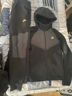 Nike svart loungewear set med huva - Nike loungewear set i svart och mörkgrått med gul logga på både byxa och hoodie. Hoodien har dragkedja, huva och fickor, medan byxorna har resår i midjan och muddar vid benslut. Perfekt för chill dagar hemma eller när du vill vara bekväm.