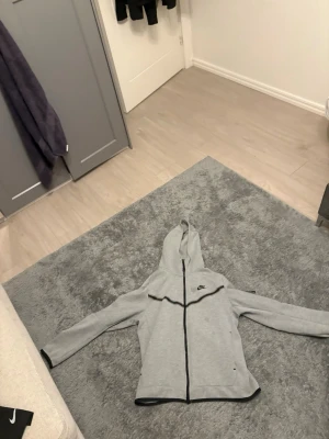 Grå zip hoodie från Nike tech  - Snygg grå hoodie från Nike med dragkedja framtill och huva. Klassisk design med svart Nike-logga på bröstet och svarta detaljer längs dragkedjan. Tillverkad i mjukt material som passar perfekt till en avslappnad stil.