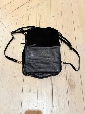 Bukvy “Bo Bardi” 5-in-1-väska i svart skinn/mocka  -  Bukvy “Bo Bardi” 5-in-1-väska i svart skinn/mocka – går att bära som ryggsäck, tote, axelremsväska, laptopväska och clutch. 