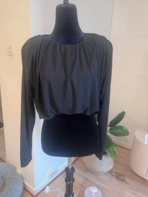 Svart croppad långärmad topp - Svart croppad topp med långa puffärmar och rund halsringning. Toppen har en lös passform och är tillverkad i ett mjukt, lätt material som faller snyggt. Perfekt för att styla med höga jeans eller kjol.