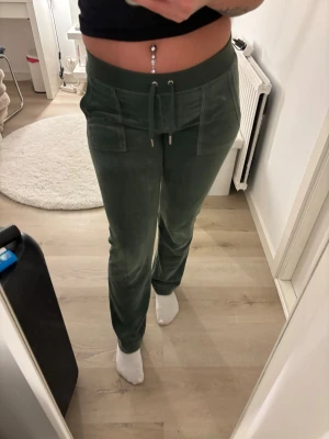 Gröna Juicy Couture med fickor - Mysiga gröna mjukisbyxor i velour med dragsko i midjan och stora fickor både fram och bak. Byxorna har raka ben och silverfärgade detaljer på bakfickorna. Perfekta för chill dagar hemma eller när du vill vara bekväm.