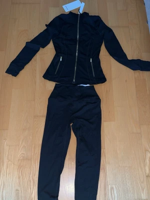Svart loungewear set med dragkedja - Lululemon Snyggt svart loungewear set med långärmad zip-tröja och matchande byxor. Tröjan har figurnära passform, dragkedja framtill och två fickor med guldfärgade dragkedjor. Byxorna är höga i midjan och har en smal siluett. Perfekt för chill dagar hemma. Storlek 2 / S