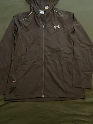 Svart hoodie från Under Armour - Snygg svart hoodie från Under Armour med dragkedja hela vägen, huva och vita loggor både fram och bak. Tillverkad i ett tekniskt material som känns sportigt och lätt. Perfekt för dig som gillar en clean och sportig stil.