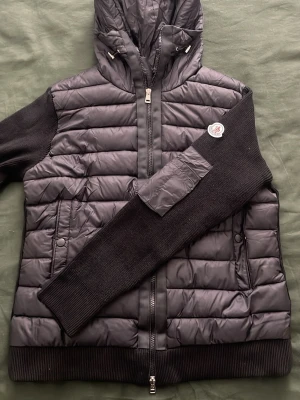 Svart moncler cardigan med scan  - Snygg svart dunjacka från Moncler med huva och dragkedja framtill. Jackan har stickade svarta ärmar och ribbade muddar, samt två fickor med knappstängning. Moncler-logga på ena ärmen och coolt serietryck på insidan. Perfekt för kalla vinterdagar.