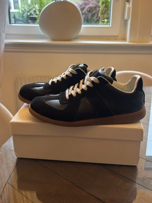 Maison margiela gats - Snygga svarta sneakers med vita skosnören och bruna gummisulor. Skorna har paneler i mocka och slätare material, samt en klassisk låg siluett. Perfekta för dig som gillar stilrena och tidlösa sneakers.