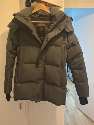 Grå Canada goose dunjacka med huva - Snygg grå Canada goose dunjacka med stor huva och quiltad design. Jackan har dragkedja och knappar framtill, två sidofickor och muddar vid ärmsluten. Perfekt för kalla vinterdagar och ger en stilren look. Pris kan diskuteras