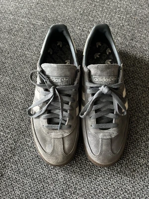 Adidas Handball Spezial grå sneakers - Säljer ett par grå Adidas Handball Spezial sneakers i mocka med klassiska vita stripes på sidorna och svart yttersula. Skorna har låga skaft och snörning framtill. Perfekta för dig som gillar retrostil och streetwear.