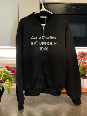 Svart zip hoodie Acne Studios - Säljer en svart zip hoodie från Acne Studios med vit text 'Acne Studios STOCKHOLM 1996' på bröstet. Tröjan har dragkedja framtill, ribbade muddar och en avslappnad passform. Perfekt för en cool och enkel streetwear-look.