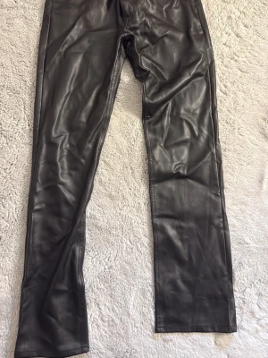 Svarta fuskskinnbyxor från BikBok - Snygga svarta byxor i fuskskinn från BikBok. De har raka ben, hög midja och bälteshällor. Materialet ger en cool och trendig look som passar perfekt till olika stilar. Stängs med knapp och dragkedja framtill.