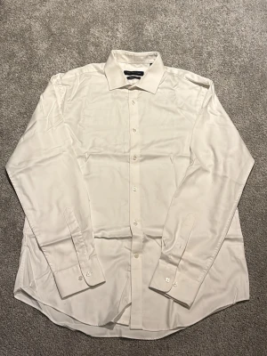 Tommy Hilfiger Vit långärmad skjorta i silke - Klassisk vit skjorta i silkesmaterial med lång ärm och knappar framtill. Skjortan har en stilren krage och manschetter med knappstängning. Perfekt för dig som gillar en clean och elegant look.