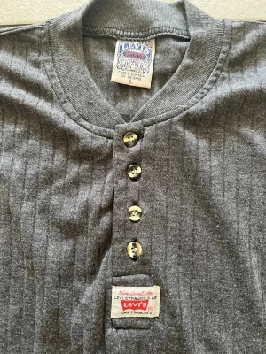 Grå ribbad topp från Levi's - Säljer en grå ribbad topp från Levi's med korta ärmar och fyra knappar framtill. Toppen har en rund halsringning och en liten Levi's-label på magen. Perfekt för en chill och avslappnad stil.