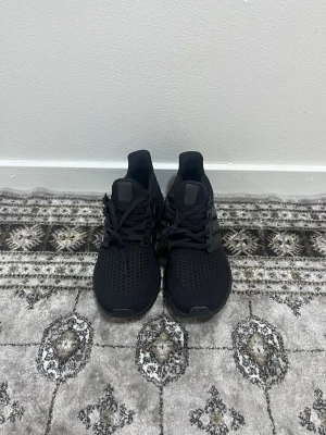 Ultraboost  - Snygga helsvarta Adidas Ultraboost sneakers med stickad ovandel och klassiska tre ränder på sidan. Skorna har en låg siluett och bekväm Boost-sula som ger bra dämpning. Perfekta för dig som vill ha stilrena och sportiga sneakers till vardags.