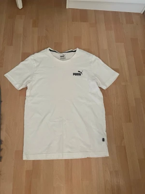 Vit t-shirt från Puma - Klassisk vit t-shirt från Puma med svart logga på bröstet. T-shirten har rund halsringning och korta ärmar. Tillverkad i mjuk bomull som känns skön mot huden. Perfekt basplagg till garderoben.