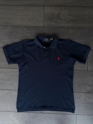 Marinblå pikétröja från Polo Ralph Lauren - Klassisk marinblå pikétröja från Polo Ralph Lauren med korta ärmar och krage. Tröjan har två knappar vid halsen och den ikoniska röda logotypen broderad på bröstet. Perfekt till jeans eller shorts för en clean look.