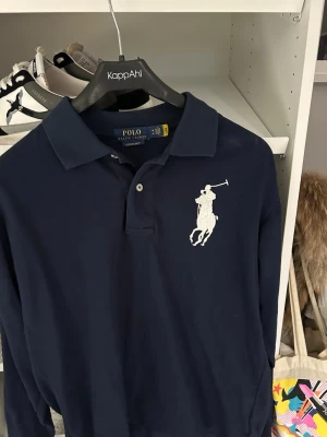 Mörkblå långärmad piké från Polo Ralph Lauren - Snygg mörkblå långärmad pikétröja från Polo Ralph Lauren med klassisk krage, knappar framtill och stort vitt polospelar-tryck på bröstet. På vänster ärm finns en vit siffra 3. Perfekt till en avslappnad men stilren look.