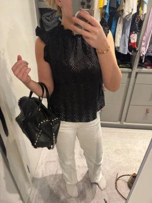 Svart ärmlös blus med volang - Säljer en svart ärmlös blus med volangdetaljer vid axlarna och halsen. Blusen har ett broderat hålmönster som ger en luftig och trendig look. Perfekt att styla med både jeans och kjol för en cool vibe.