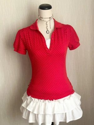 Röd prickig topp med krage - Snygg röd topp med små vita prickar och klassisk krage. Modellen är figurnära med korta ärmar och v-ringad hals. Perfekt för dig som gillar retrovibbar och vill sticka ut lite extra. Matcha med kjol eller jeans för en cool look.