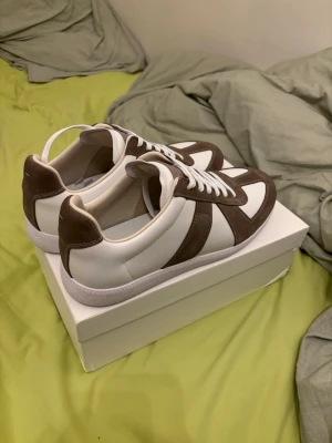 Bruna och vita sneakers i mocka och skinn - Snygga sneakers i vitt skinn med bruna mockadetaljer och vita snören. Klassisk låg modell med stilren design och bekväm sula. Perfekta för dig som gillar en clean och tidlös look. Finns ett litet hål på sidan av skon 