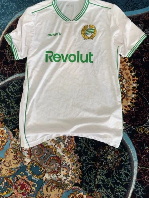Vit fotbollströja Hammarby x Craft - Säljer en vit fotbollströja från Hammarby i samarbete med Craft. Tröjan har gröna detaljer vid krage och ärmslut, samt klubbmärke och sponsortryck framtill. Tillverkad i polyester och har korta ärmar. Perfekt för dig som älskar fotboll och vill stötta laget.