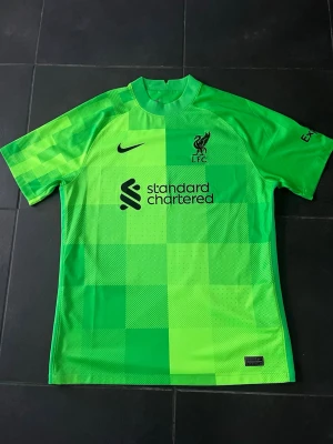Grön Liverpool målvaktströja Nike - Säljer en grön Liverpool FC målvaktströja från Nike med svart logga och klubbmärke på bröstet. Tröjan har ett rutmönster framtill, korta ärmar och rund hals. Tillverkad i lätt och ventilerande material, perfekt för fotbollsträning eller match.