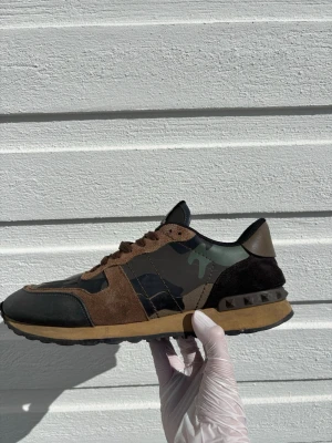 Valentino rockrunners  - Snygga sneakers med kamouflagemönster i grönt och svart, kombinerat med bruna mockadetaljer och svarta partier. Skon har en låg siluett och coola nitar på hälen. Materialmix av mocka, läder och textil ger en unik look.