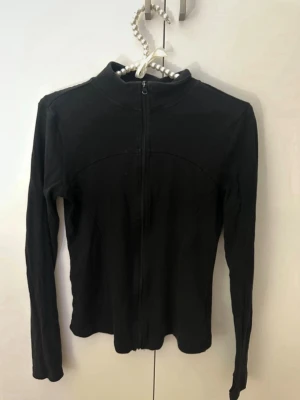 Svart stickad ziptröja - Säljer en svart stickad ziptröja med lång ärm och hög krage. Tröjan har hel dragkedja framtill och en enkel, stilren design. Perfekt att slänga på sig över en t-shirt för en clean look.