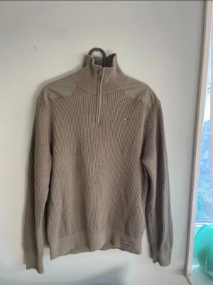 Beige stickad tröja Tommy Hilfiger - Stickad tröja i grön från Tommy Hilfiger med halv dragkedja och ribbade muddar. Tröjan har förstärkta axelpartier och det klassiska Hilfiger-logotypen broderad på bröstet. Grovstickad struktur ger en avslappnad och trendig look.