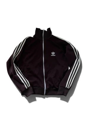 Brun Adidas track jacket med vita ränder - Säljer en brun Adidas track jacket med klassiska vita ränder längs ärmarna och vit dragkedja. Jackan har hög krage, muddar vid ärmslut och nederkant samt Adidas logga på bröstet. Perfekt för en sportig och retro look.
