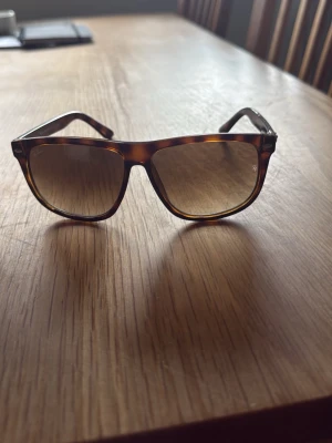 Bruna solglasögon från Ray-Ban - Snygga solglasögon från Ray-Ban med klassisk fyrkantig form och brunmelerad båge. Glasen är tonade i brunt och bågarna har en skön retrovibe. Perfekta för soliga dagar och ger en cool look till vilken outfit som helst.