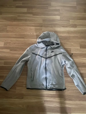 Grå Nike hoodie med dragkedja - Snygg grå hoodie från Nike med svart dragkedja och svarta detaljer. Hoodien har huva, långa ärmar och två fickor framtill. Perfekt för en sportig och avslappnad stil. Nike-logga på bröstet ger en klassisk look.