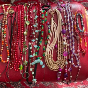 Färgglada halsband och armband mix - Samling med 23 st. unika halsband och armband i olika färger och former. Här finns pärlor i rött, grönt, lila, svart, orange och beige, samt detaljer i guld och silver. Materialen varierar mellan plast, metall och trä. Perfekt för dig som gillar att mixa och matcha smycken med olika uttryck.
