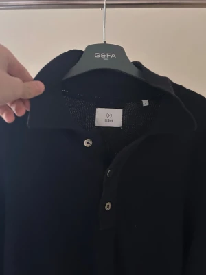 Svart cardigan/kofta med krage - Klassisk svart stickad tröja från BiBch med krage och knäppning framtill. Tröjan har långa ärmar och ribbade muddar vid ärmslut och nederkant. Perfekt för en stilren och enkel look.