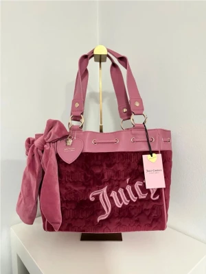 Rosa sammetshandväska Juicy Couture - Superfin rosa handväska från Juicy Couture i sammet och skinnimitation. Väskan har dubbla handtag, dekorativ rosett och hjärtformad tag. Broderad logga framtill och snygga metalldetaljer. Perfekt för dig som gillar en trendig och söt look.