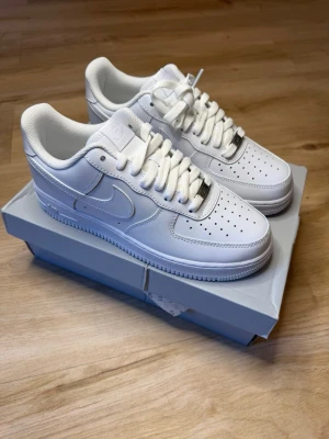 Vita Nike Air Force 1 sneakers - Klassiska helvita Nike Air Force 1 sneakers i lågt utförande. Skorna har en stilren design med perforerad tå, vita skosnören och den ikoniska swoosh-loggan på sidan. Tillverkade i läder med gummisula för skön komfort och streetstyle.