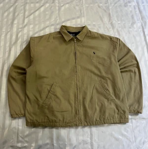 Beige casual jacka med dragkedja - Snygg beige jacka med dragkedja framtill och två snedställda fickor. Klassisk krage och minimalistisk design med liten broderad logga på bröstet. Perfekt för dig som gillar enkel och stilren look. Materialet ser ut att vara bomull.
