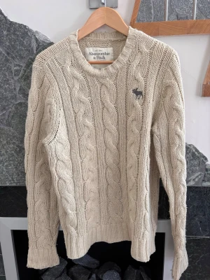 Beige kabelstickad tröja Abercrombie - Kabelstickad tröja i beige från Abercrombie & Fitch. Tröjan har rund halsringning, långa ärmar och en liten mörk logga med älg på bröstet. Tillverkad i mjukt, tjockt material som ger en klassisk och avslappnad look.