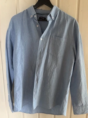 Ljusblå skjorta från In Our Terms - Klassisk ljusblå skjorta från In Our Terms med button-down krage och bröstficka. Skjortan har långa ärmar och vita knappar framtill. Perfekt till jeans eller chinos för en clean och enkel stil.