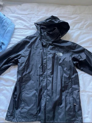 Svart regnjacka från The North Face - Snygg svart regnjacka med huva och dragkedja framtill. Jackan har två sidofickor med dragkedja och ett foder som känns mjukt mot kroppen. Perfekt för regniga dagar och outdoor-äventyr. Står L men är en S, ej äkta men bra kvalitet 