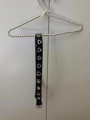 Svart bälte med hjärtan i silver - Snyggt svart bälte med silverfärgade hjärtformade hål längs hela bandet. Perfekt accessoar för att ge din outfit en edgy och unik touch. Bältet har ett klassiskt spänne i silver och är gjort i syntetmaterial.