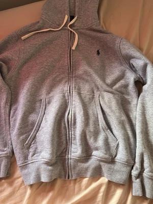 Grå zip-hoodie från Polo Ralph Lauren - Snygg grå hoodie med dragkedja från Polo Ralph Lauren. Klassisk design med huva och vita snören, två fickor framtill och broderad logga på bröstet. Perfekt för en chill och stilren look.