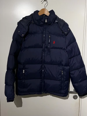 Mörkblå pufferjacka med huva - Snygg mörkblå pufferjacka med avtagbar huva och dragkedja framtill. Jackan har flera fickor med dragkedja och en röd broderad logga på bröstet. Perfekt för kalla dagar och riktigt skön att bära.