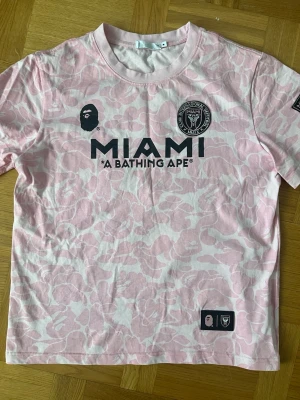Inter Miami Bape tröja - Säljer en rosa och vit camo t-shirt från samarbetet MIAMI x A Bathing Ape. T-shirten har korta ärmar, rund hals och tryck framtill med MIAMI-logga och BAPE-apa. Perfekt för dig som gillar streetwear och fotboll.